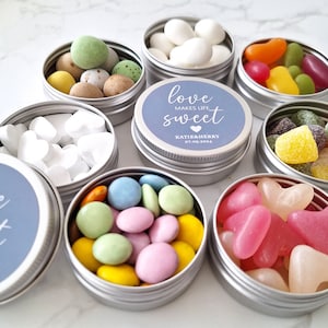 May include: Small silver tins filled with colorful candies, including jelly beans, heart-shaped sweets, and sugared almonds. Some tins have blue labels with the text "love makes life sweet" and a date. A variety of treats are visible.