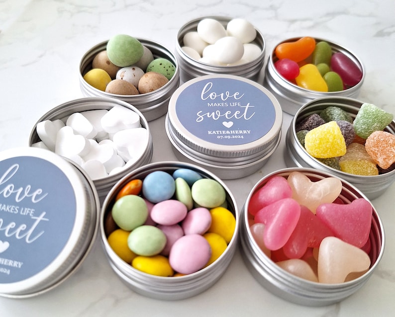May include: Small silver tins filled with colorful candies, including jelly beans, heart-shaped sweets, and sugared almonds. Some tins have blue labels with the text "love makes life sweet" and a date. A variety of treats are visible.