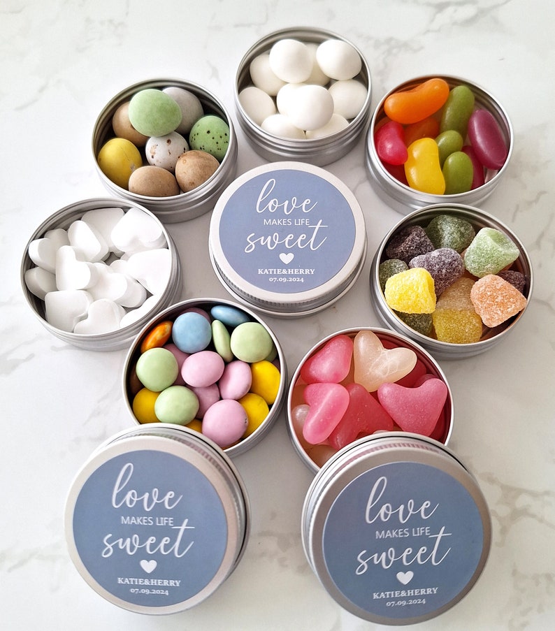 May include: A collection of small, round, silver tins filled with various candies, including white heart-shaped mints, colorful jelly beans, and chocolate-covered candies. Some tins have blue lids with the text "love makes life sweet".