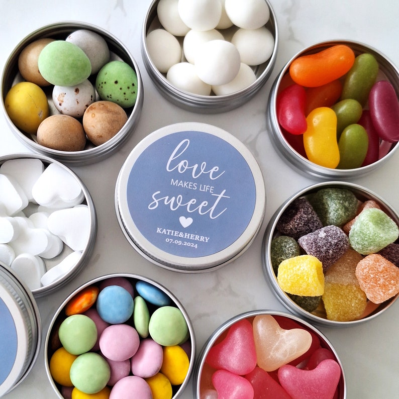 May include: Assortment of candies in small, round, silver tins. The tins contain various sweets, including jelly beans, heart-shaped candies, and chocolate-covered treats. A central tin has a blue label with the text "love makes life sweet".