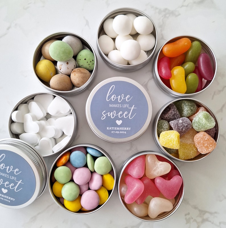 May include: An overhead shot showcases several small, round, silver tins filled with colorful candies. Some tins contain heart-shaped candies, while others hold jelly beans, pastel-colored chocolates, and sugared almonds. The central tin has a blue label with the text "love makes life sweet".
