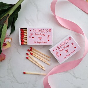 Op de afbeelding: Twee rechthoekige luciferdoosjes met lichtroze etiketten met rode tekst die OLIVIA'S Hen Party en The Perfect Match leest. De etiketten bevatten rode illustraties van een champagnefles, een ring, een strik, hartjes en een discobal. Lucifers met rode koppen.