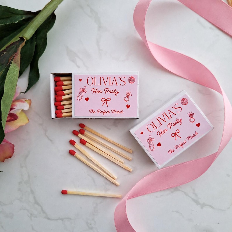 Op de afbeelding: Twee rechthoekige luciferdoosjes met lichtroze etiketten met rode tekst die OLIVIA'S Hen Party en The Perfect Match leest. De etiketten bevatten rode illustraties van een champagnefles, een ring, een strik, hartjes en een discobal. Lucifers met rode koppen.