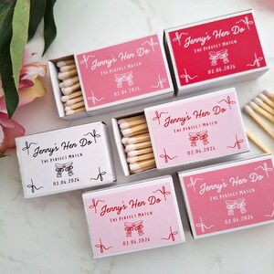 Op de afbeelding: Een verzameling rechthoekige luciferdoosjes in roze, rode en witte tinten. Elk doosje bevat de tekst Jenny's Hen Do, The Perfect Match, een afbeelding van twee gestrikte linten en de datum 02.06.2026. Sommige doosjes zijn geopend.
