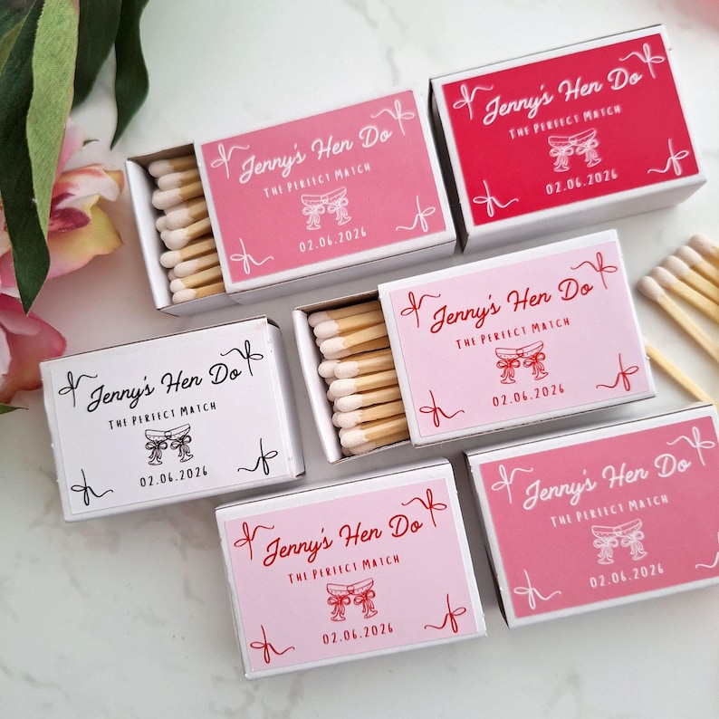 Op de afbeelding: Een verzameling rechthoekige luciferdoosjes in roze, rode en witte tinten. Elk doosje bevat de tekst Jenny's Hen Do, The Perfect Match, een afbeelding van twee gestrikte linten en de datum 02.06.2026. Sommige doosjes zijn geopend.