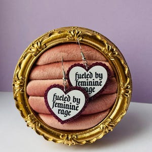 Könnte beinhalten: Herzförmige Ohrringe mit dem Text "fueled by feminine rage" in gotischer Schrift. Die Ohrringe haben einen weißen Hintergrund mit einem dunkelroten Rand und werden in einem goldgerahmten, samtgefütterten Display präsentiert.