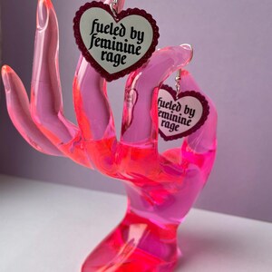 May include: A pair of heart-shaped earrings with the text "fueled by feminine rage" displayed on a white background. The earrings are held by a pink, translucent hand-shaped display stand.