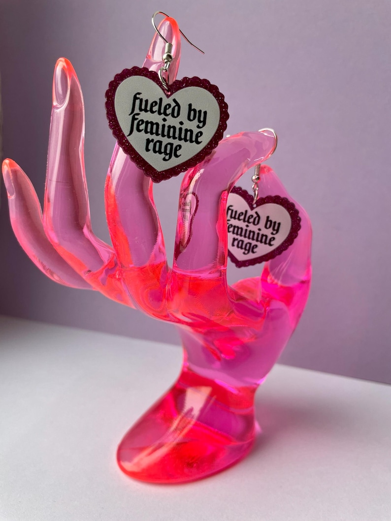 May include: A pair of heart-shaped earrings with the text "fueled by feminine rage" displayed on a white background. The earrings are held by a pink, translucent hand-shaped display stand.