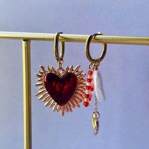Peut inclure: Boucles d'oreilles créoles dorées. L'une présente un pendentif cœur rouge avec un motif de rayons de soleil. L'autre comporte des perles rouges et roses, une perle blanche et un petit charme doré.