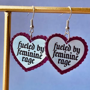 Könnte beinhalten: Ein Paar herzförmige Ohrringe mit weißer Mitte und dunkelrosa Glitzerbordüre. Der Text "fueled by feminine rage" steht in schwarzer Schrift auf den Ohrringen. Sie hängen an silberfarbenen Ohrhaken.
