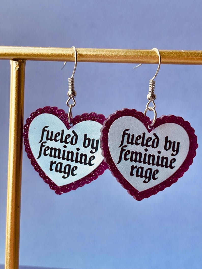 May include: A pair of heart-shaped earrings with a white center and a dark pink glittery border. The earrings have the text "fueled by feminine rage" in black script. The earrings hang from silver-colored fish hook ear wires.