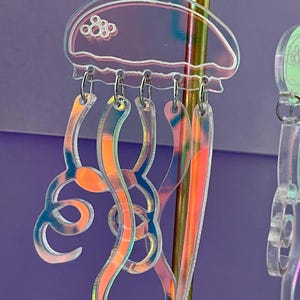 May include: Iridescent jellyfish earrings with orange, pink, and blue accents. The earrings feature a clear, translucent design with a jellyfish shape, including a bell and flowing tentacles. The earrings are suspended from silver-colored hooks.