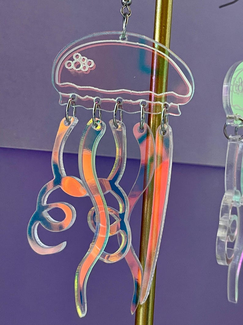 May include: Iridescent jellyfish earrings with orange, pink, and blue accents. The earrings feature a clear, translucent design with a jellyfish shape, including a bell and flowing tentacles. The earrings are suspended from silver-colored hooks.