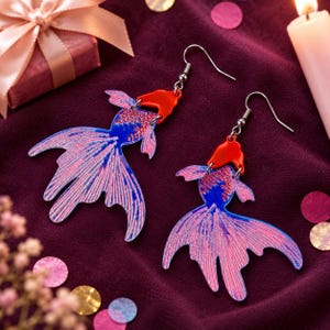 May include: A pair of dangle earrings shaped like goldfish, featuring vibrant blue bodies with intricate pink scale patterns and bright red heads. The earrings are attached to silver-toned metal hooks, showcasing a detailed fish silhouette design.
