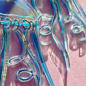 May include: Close-up of iridescent blue earrings with intricate, swirling designs. The earrings feature multiple layers and textures, with a mix of clear and colored elements. The design includes loops and dangling strands, creating a unique and eye-catching accessory.