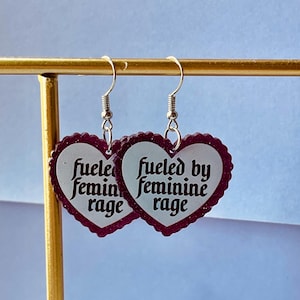 May include: A pair of heart-shaped earrings with a scalloped edge and the text "fueled by feminine rage" in a vintage font. The earrings are white with a dark red border and silver hooks.