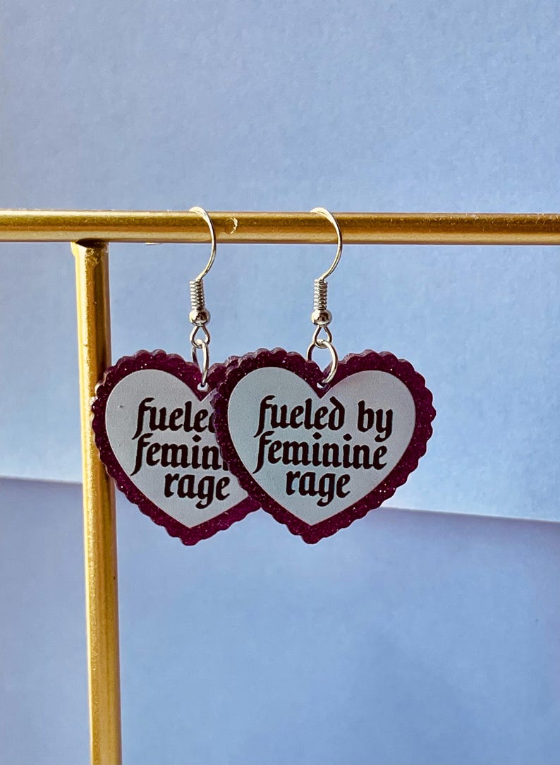 May include: A pair of heart-shaped earrings with a scalloped edge and the text "fueled by feminine rage" in a vintage font. The earrings are white with a dark red border and silver hooks.