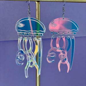 Könnte beinhalten: Ein Paar schillernde Quallenohrringe. Jeder Ohrring hat eine blaue oder rosa Kuppel mit weißem Rand und hängenden, klaren, gebogenen Tentakeln. Die Ohrringe hängen an silberfarbenen Fischhaken-Ohrdrähten. Der Hintergrund ist einfarbig lila.