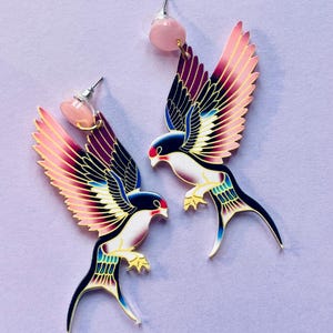 Puede incluir: Un par de pendientes con forma de pájaro. Los pendientes presentan un diseño colorido con acentos rosas, morados, azules y dorados. Cada pendiente tiene una cuenta rosa en la parte superior y una cola larga y curvada. Los pájaros se enfrentan.