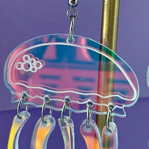 May include: Iridescent jellyfish earrings with a clear, pink, and blue design. The earrings have a jellyfish shape with dangling, curved tentacles. The jellyfish top has a white outline and a cluster of white circles. The earrings hang from silver-colored hooks.