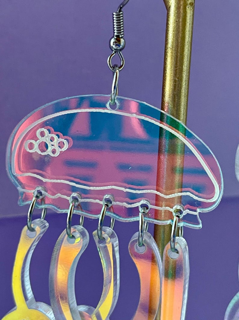 May include: Iridescent jellyfish earrings with a clear, pink, and blue design. The earrings have a jellyfish shape with dangling, curved tentacles. The jellyfish top has a white outline and a cluster of white circles. The earrings hang from silver-colored hooks.