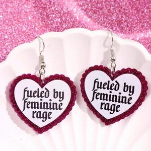 May include: A pair of heart-shaped earrings with a white center and a glittery burgundy border. The earrings have the text "fueled by feminine rage" in black script. The earrings hang from silver-colored hooks.