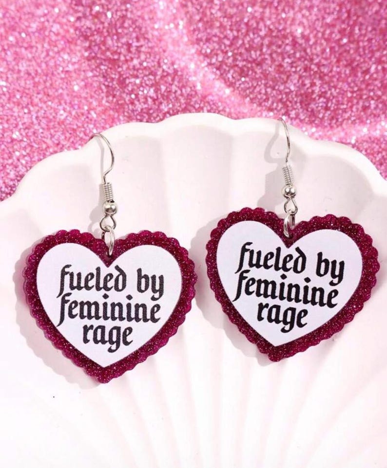 May include: A pair of heart-shaped earrings with a white center and a glittery burgundy border. The earrings have the text "fueled by feminine rage" in black script. The earrings hang from silver-colored hooks.