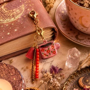 Könnte beinhalten: Ein goldfarbener Metallanhänger mit einer rosa Tasse mit Blumenmuster, einer kleinen rosa Quaste, einer Reihe bronzefarbener Metallperlen und einer Reihe leuchtend orangefarbener Perlen, befestigt an einem goldenen Karabinerverschluss.