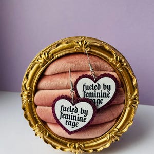 May include: A pair of heart-shaped earrings with the text "fueled by feminine rage" in a white font. The earrings have a dark red glitter border and are displayed in a gold-colored ornate frame.