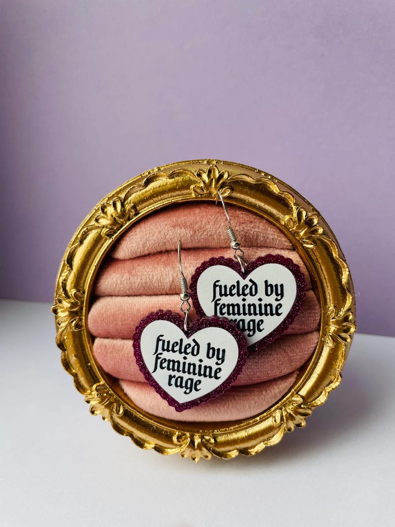 May include: A pair of heart-shaped earrings with the text "fueled by feminine rage" in a white font. The earrings have a dark red glitter border and are displayed in a gold-colored ornate frame.