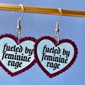 May include: A pair of heart-shaped earrings with a light blue center and a dark purple glittery border. The earrings have the text "fueled by feminine rage" in black script. The earrings hang from silver-colored fish hook ear wires.