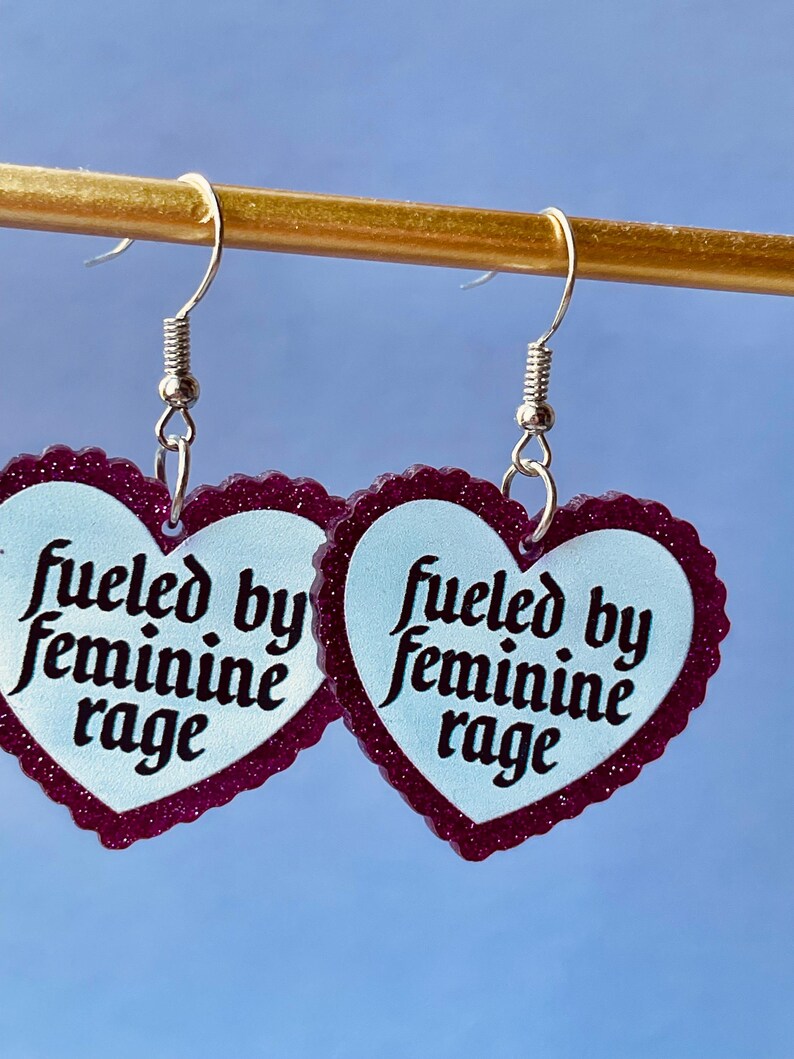 May include: A pair of heart-shaped earrings with a light blue center and a dark purple glittery border. The earrings have the text "fueled by feminine rage" in black script. The earrings hang from silver-colored fish hook ear wires.