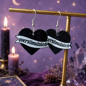 May include: A pair of heart-shaped dangle earrings featuring a black glitter finish and a white banner across the center with the word "overstimulated" printed in black text. The earrings hang from silver-toned metal hooks on a gold-toned stand.