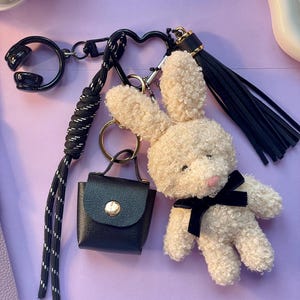 Könnte beinhalten: Ein Schlüsselanhänger mit einem cremefarbenen Plüschhasen mit schwarzer Schleife, einer kleinen schwarzen Handtasche und einem schwarzen Quastenanhänger. Der Schlüsselanhänger enthält einen schwarzen herzförmigen Verschluss und ein schwarz-weiß gemustertes Schlüsselband.