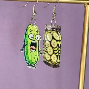Könnte beinhalten: Ein Paar ausgefallene Ohrhänger mit Cartoon-Gurken-Motiven. Ein Ohrring zeigt eine grüne Gurke mit überraschtem Gesichtsausdruck, der andere ein Glas Gurken. Sie hängen an silberfarbenen Fischhaken-Ohrdrähten. Ein lustiges Accessoire.