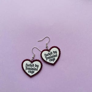 May include: A pair of heart-shaped earrings with the text "fueled by feminine rage" in black script. The earrings have a white background with a dark red glittery border and silver hooks, set against a light purple backdrop.