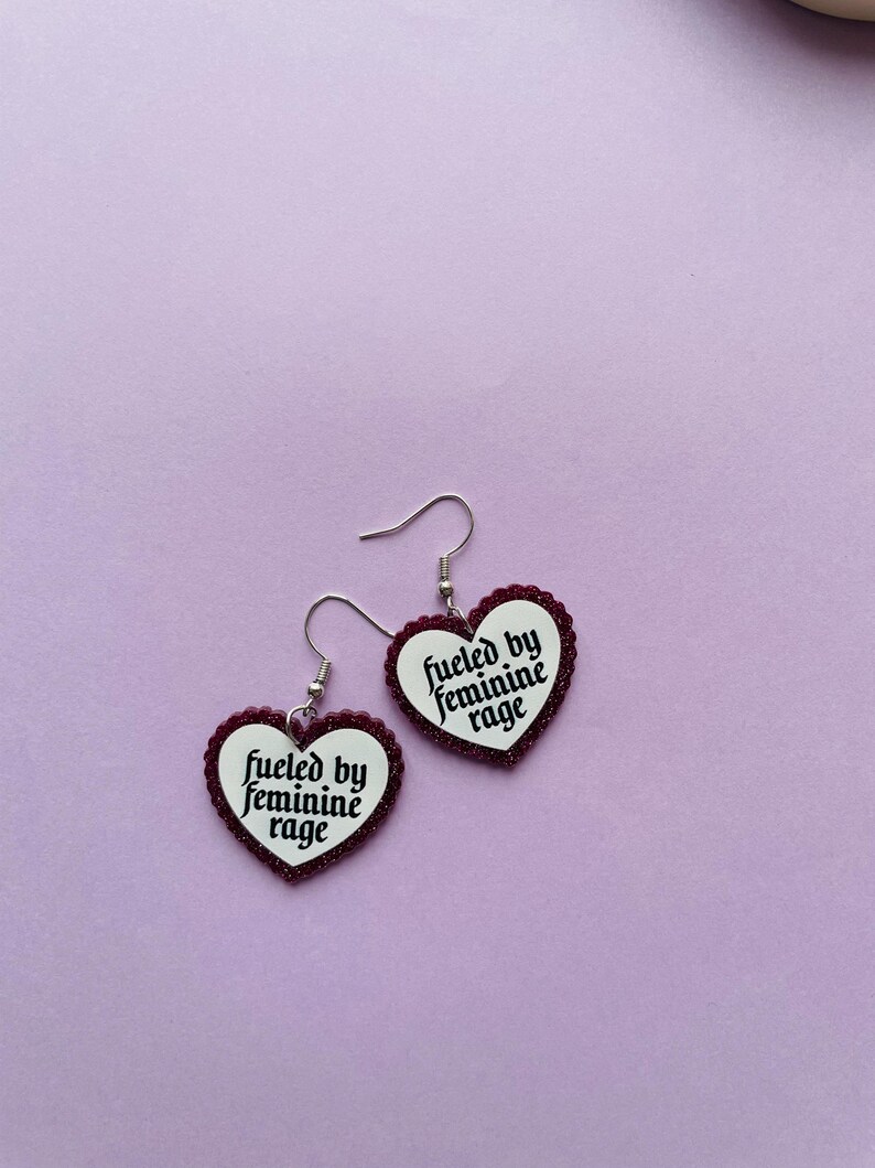 May include: A pair of heart-shaped earrings with the text "fueled by feminine rage" in black script. The earrings have a white background with a dark red glittery border and silver hooks, set against a light purple backdrop.