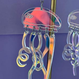 May include: Iridescent jellyfish earrings with a clear, holographic design. The earrings feature a jellyfish shape with a rounded top and flowing tentacles. The earrings are suspended from silver-colored ear wires.