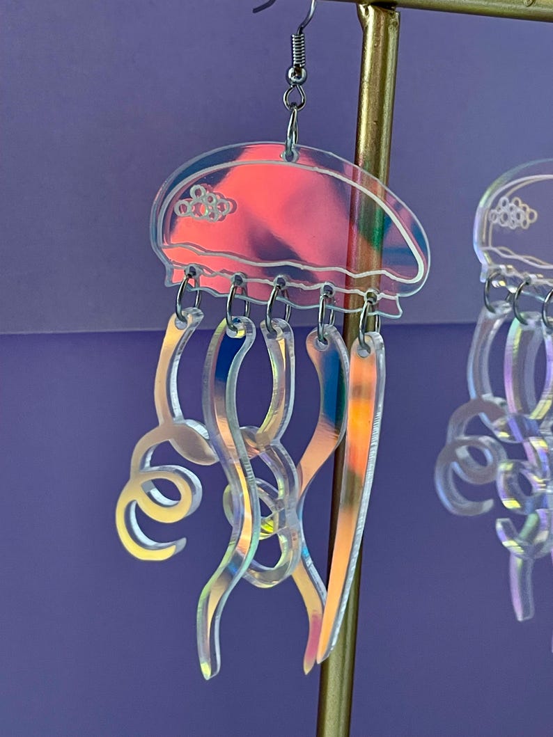 May include: Iridescent jellyfish earrings with a clear, holographic design. The earrings feature a jellyfish shape with a rounded top and flowing tentacles. The earrings are suspended from silver-colored ear wires.