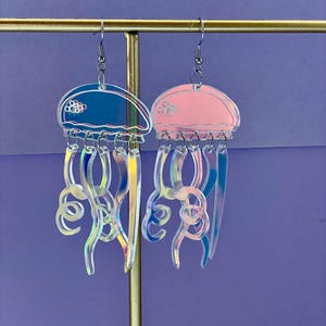 May include: A pair of jellyfish earrings. One earring is blue, the other is pink. Both earrings have clear, iridescent tentacles and silver-colored ear wires. The earrings are displayed on a gold-colored metal stand against a purple background.