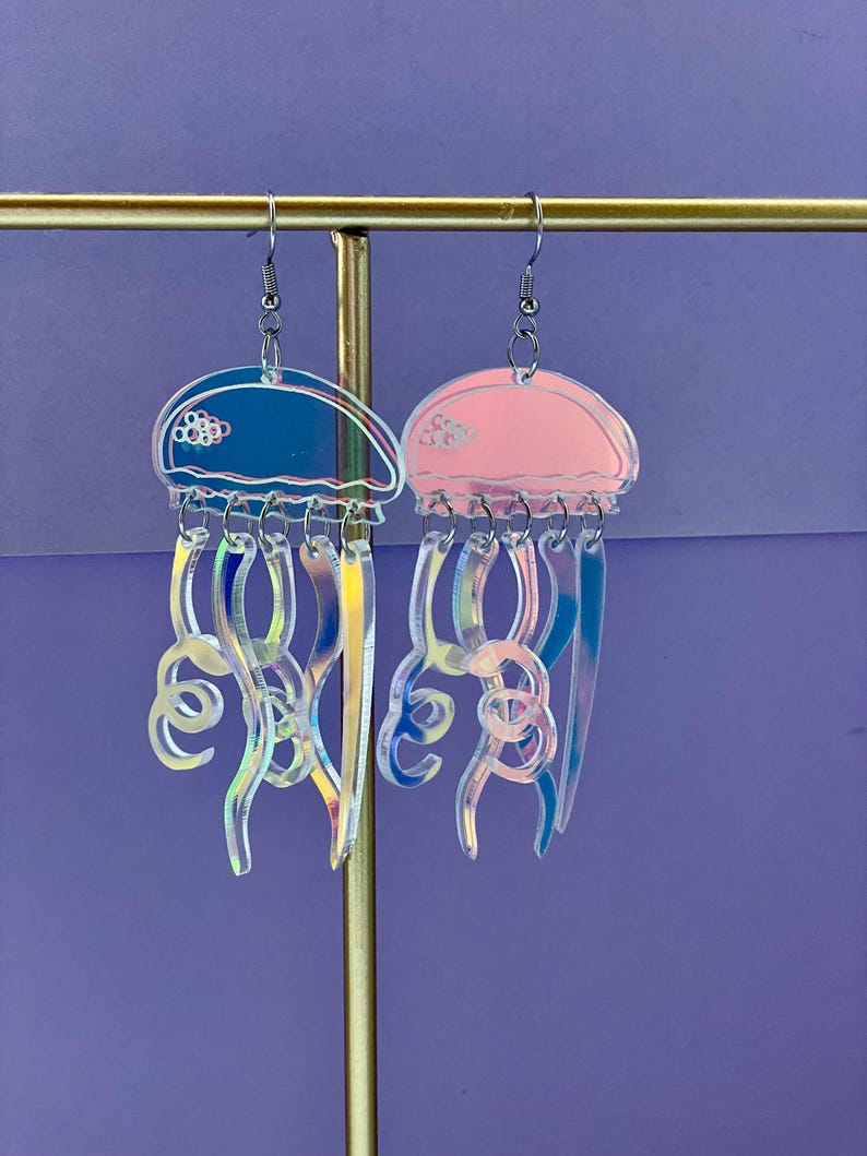 May include: A pair of jellyfish earrings. One earring is blue, the other is pink. Both earrings have clear, iridescent tentacles and silver-colored ear wires. The earrings are displayed on a gold-colored metal stand against a purple background.
