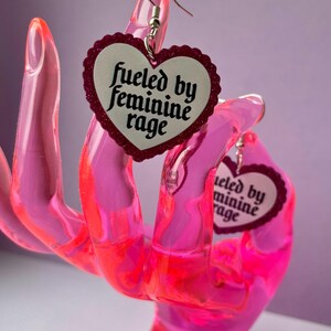 May include: A pair of heart-shaped earrings with a white center and a glittery red border. The earrings have the text "fueled by feminine rage" in black script. The earrings are displayed on a pink hand-shaped display.
