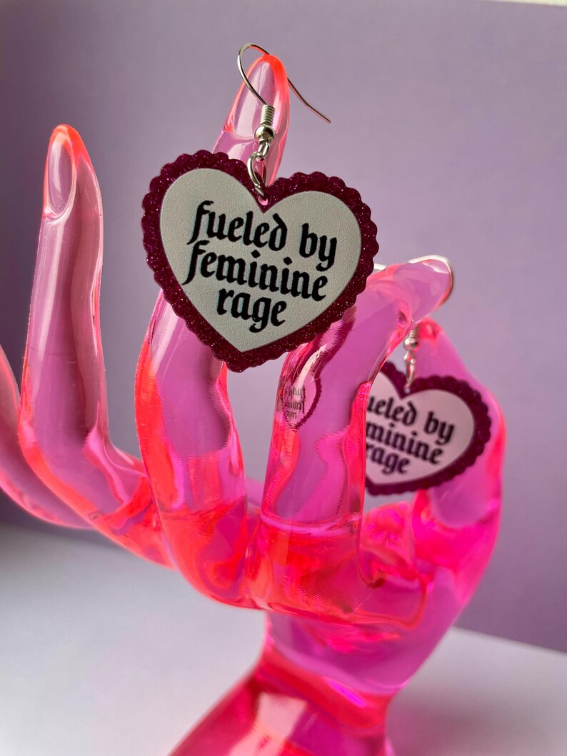 May include: A pair of heart-shaped earrings with a white center and a glittery red border. The earrings have the text "fueled by feminine rage" in black script. The earrings are displayed on a pink hand-shaped display.