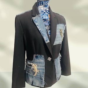 May include: A black blazer featuring blue denim patchwork panels on the lapel, chest, and lower front. The garment includes a single flower-shaped button closure and two gold-toned lizard-shaped embellishments attached to the denim sections.