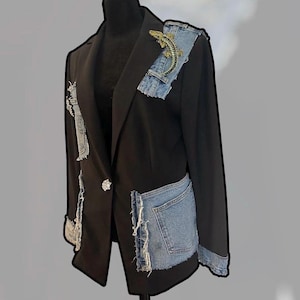 May include: A black blazer featuring light blue denim patches with frayed edges on the lapel, sleeves, and pockets. A gold-toned alligator brooch is pinned to the left lapel, and a single decorative silver-toned button fastens the front closure.