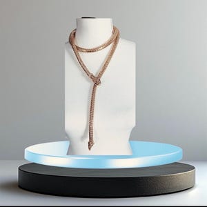 Könnte beinhalten: Eine schlangenförmige Lariat-Halskette in Roségold mit einem strukturierten, flexiblen, netzartigen Körper. Die Kette schlingt sich um den Hals und hängt in einem langen, schmalen Schwanz herab, mit einem detaillierten Schlangenkopf am tiefsten Punkt.
