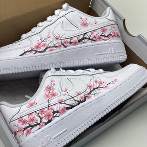 Peut inclure: Une paire de baskets blanches avec un motif peint à la main de fleurs de cerisier roses sur des branches noires s'étendant sur les côtés. Les chaussures ont des lacets blancs, une semelle intermédiaire blanche avec le mot AIR et sont dans une boîte.