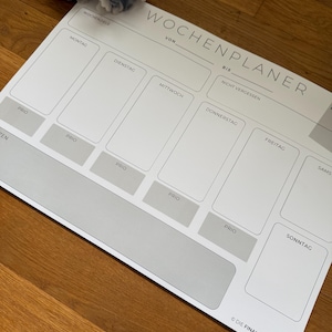 Könnte beinhalten: Ein weißer Wochenplaner mit grauen Texten und Kästchen für jeden Wochentag. Der Planer enthält den Text "WOCHENPLANER" und "DIE FINANZKOMPLIZIN". Eine dekorative Blume befindet sich in der oberen linken Ecke.