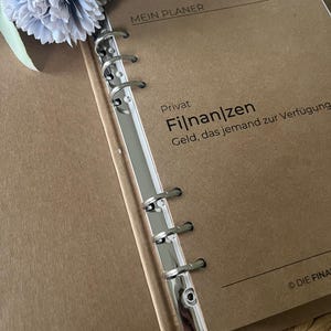 K&ouml;nnte beinhalten: Ein brauner Planer mit einem silbernen Ringbinder. Der Einband hat den Text "Mein Planer" und "Privat Finanzen Geld, das jemand zur Verf&uuml;gung." Der Planer liegt auf einer gewebten Oberfl&auml;che.