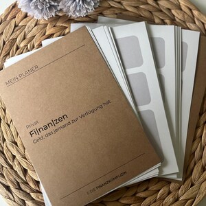 K&ouml;nnte beinhalten: Ein braunes Notizbuch mit dem Text "MEIN PLANER" und "Privat Finanzen Geld, das jemand zur Verf&uuml;gung hat" auf dem Einband. Das Notizbuch liegt auf einer geflochtenen Weideunterlage mit zwei grauen Blumen.